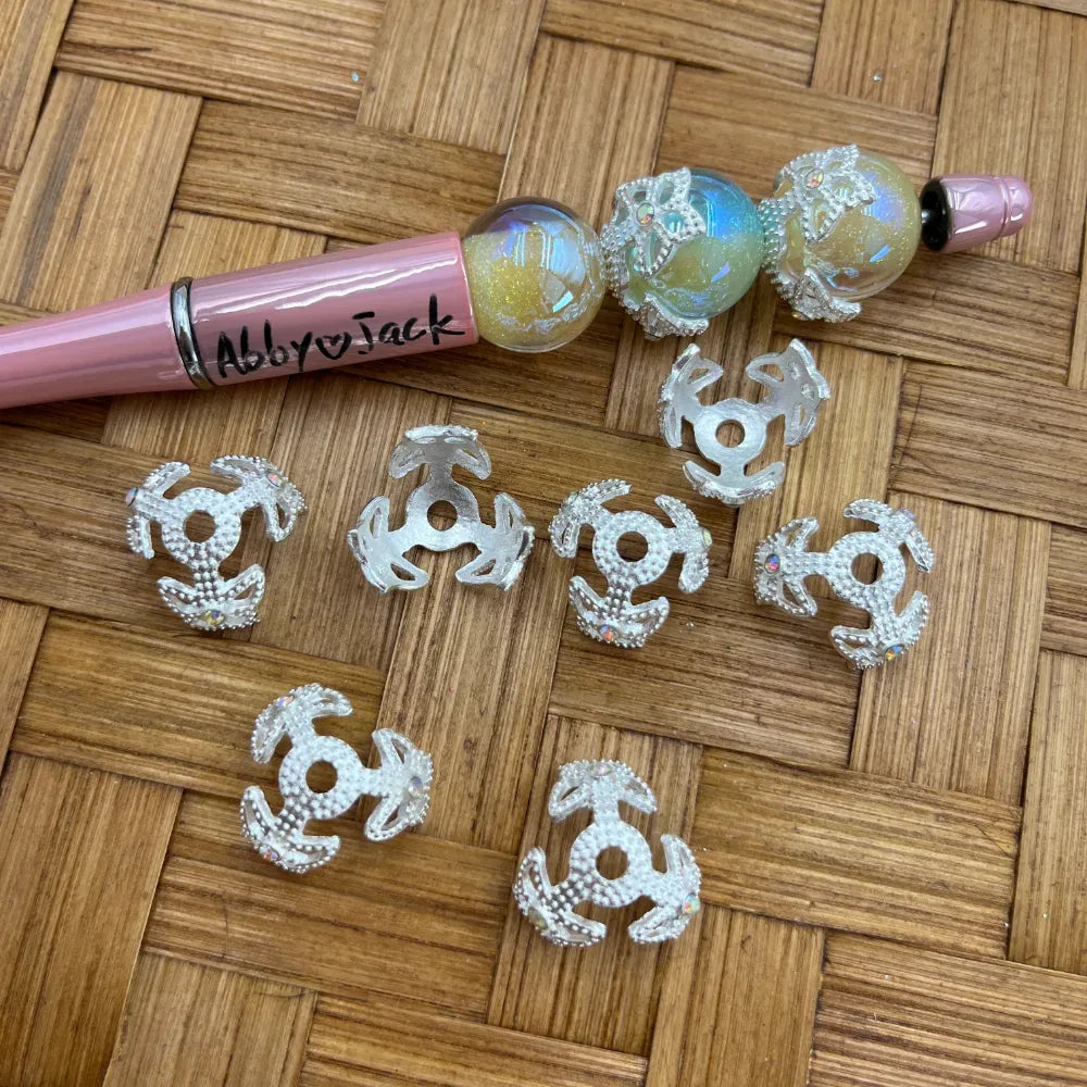 Silver filigree bead caps with iridescent AB rhinestone accents displayed on a woven surface alongside a pink beadable pen decorated with aurora borealis glitter beads, from DIY Mini Planet craft supplies.