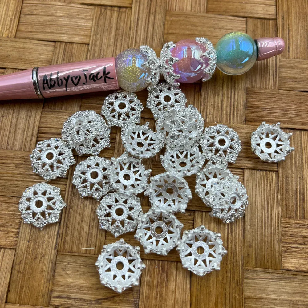 Silver filigree crown-style metal bead caps with rhinestone accents scattered on woven surface next to beadable pens with iridescent globe beads, showing intricate lattice and sunburst cutout design.