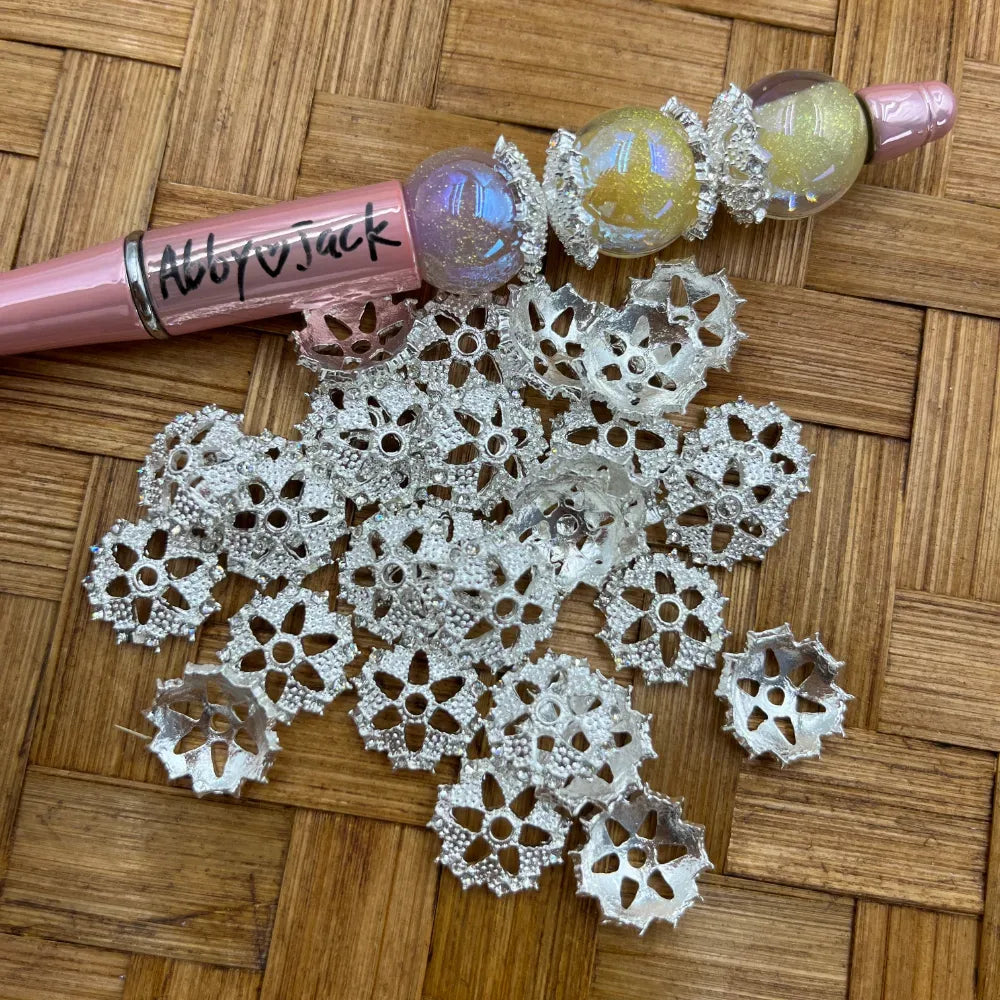 Silver filigree flower-shaped bead cap metal spacers piled on a woven bamboo surface next to a pink beadable pen decorated with iridescent glitter beads and silver bead caps.