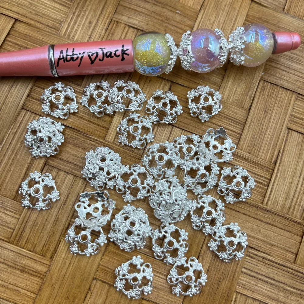 Silver filigree flower bead caps scattered on woven surface next to a beadable pen with iridescent beads, showing ornate lacy petal design used in DIY jewelry making.