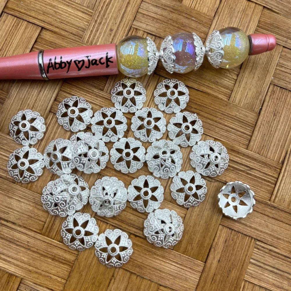 Silver filigree flower-shaped bead caps with lace cutout petal design displayed on woven bamboo surface next to a beadable pen with iridescent globe beads.