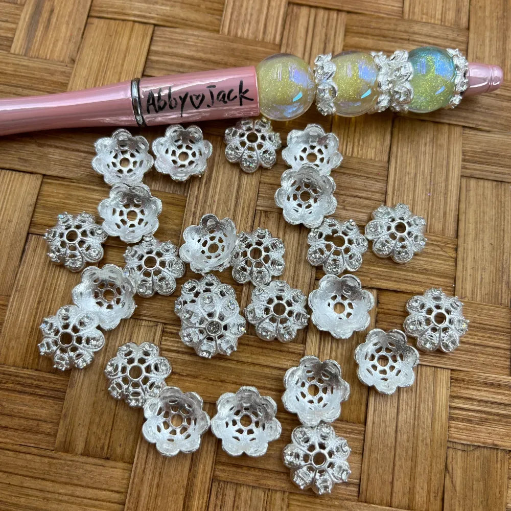 Silver filigree flower-shaped bead caps with lacy cutout petal design scattered on wicker surface next to a beadable pen with iridescent beads, by DIY Mini Planet.