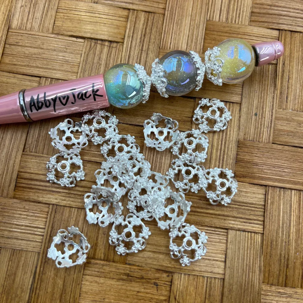 Silver filigree flower bead cap metal spacers scattered on wicker surface next to a pink beadable pen decorated with iridescent glitter globe beads and silver bead caps.