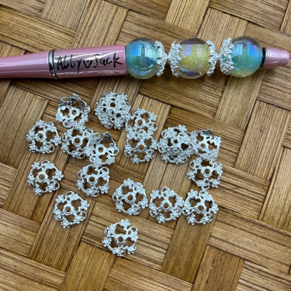 Silver filigree flower bead caps spacers displayed on woven bamboo surface next to a pink beadable pen decorated with iridescent globe beads and matching silver bead caps.