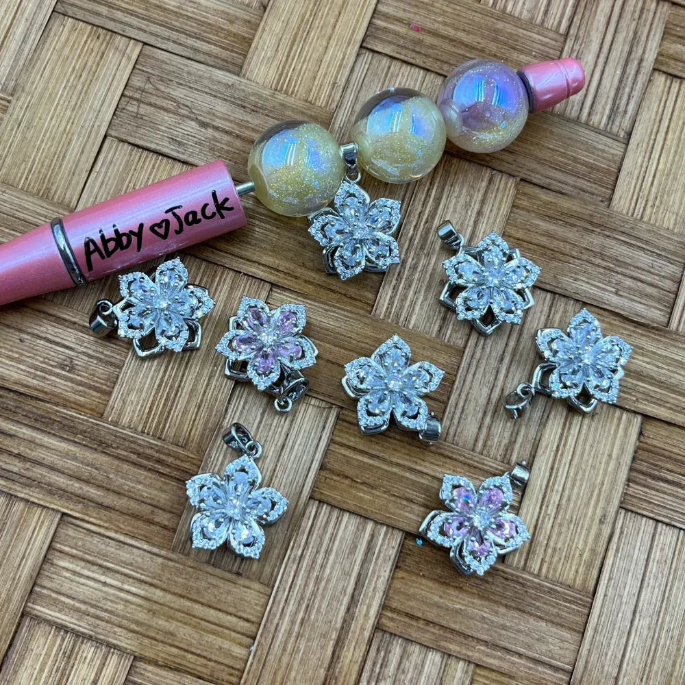 Silver flower-shaped bail beads with clear and pink cubic zirconia crystals arranged on a woven surface next to iridescent globe beads and a beadable pen, for DIY jewelry making and beadable pens.
