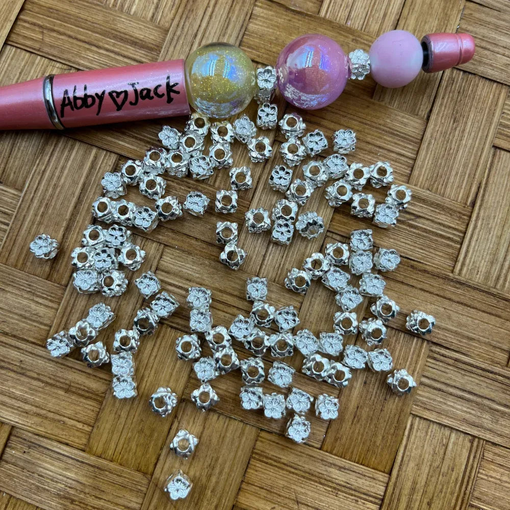 Silver flower daisy patterned metal spacer beads with large holes scattered on a woven surface next to beadable pens, used for DIY jewelry making, bracelets, and keychains.