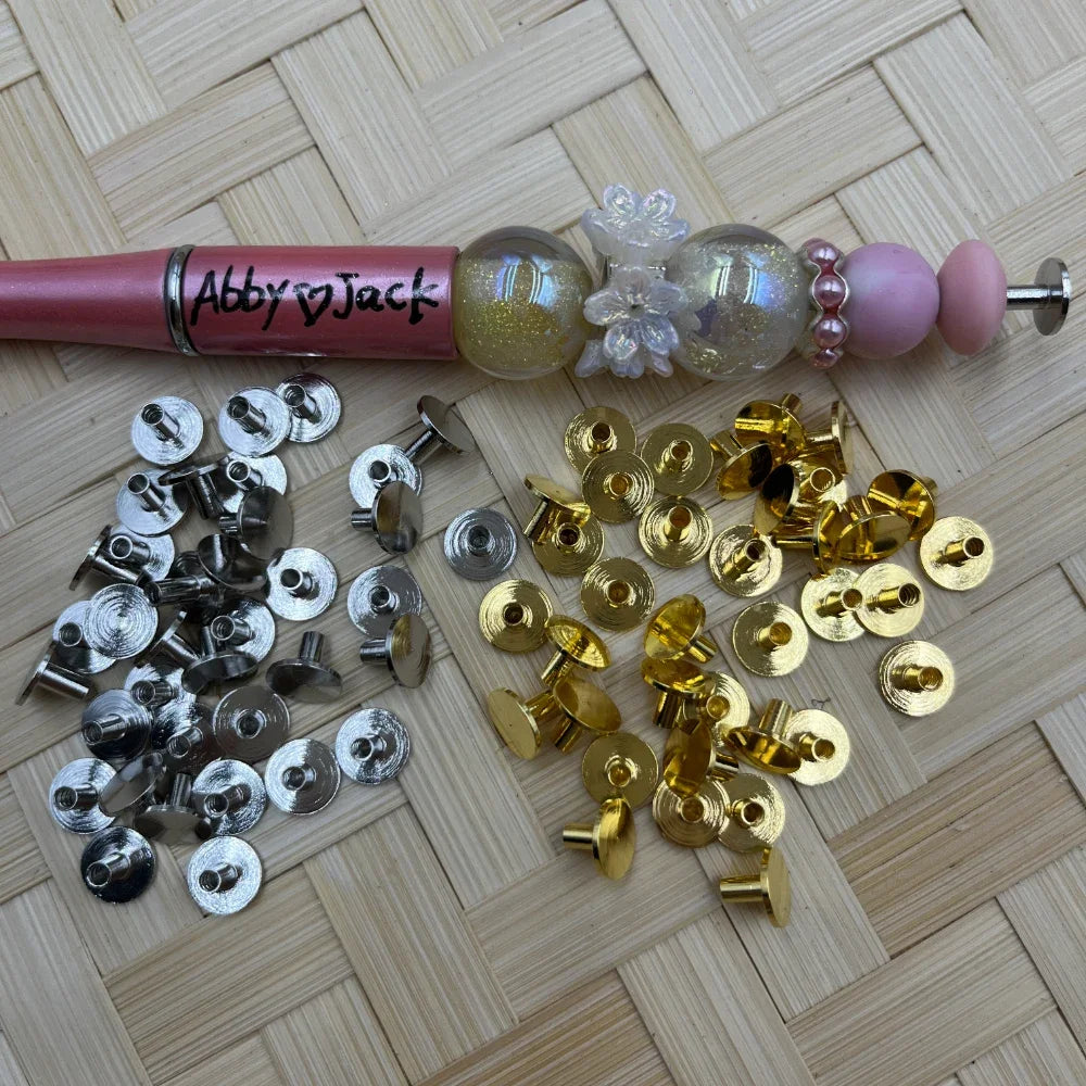 Silver and gold metal rivet end cap pen toppers displayed next to a decorated beadable pen with glitter globe and pink pearl beads on a woven surface.