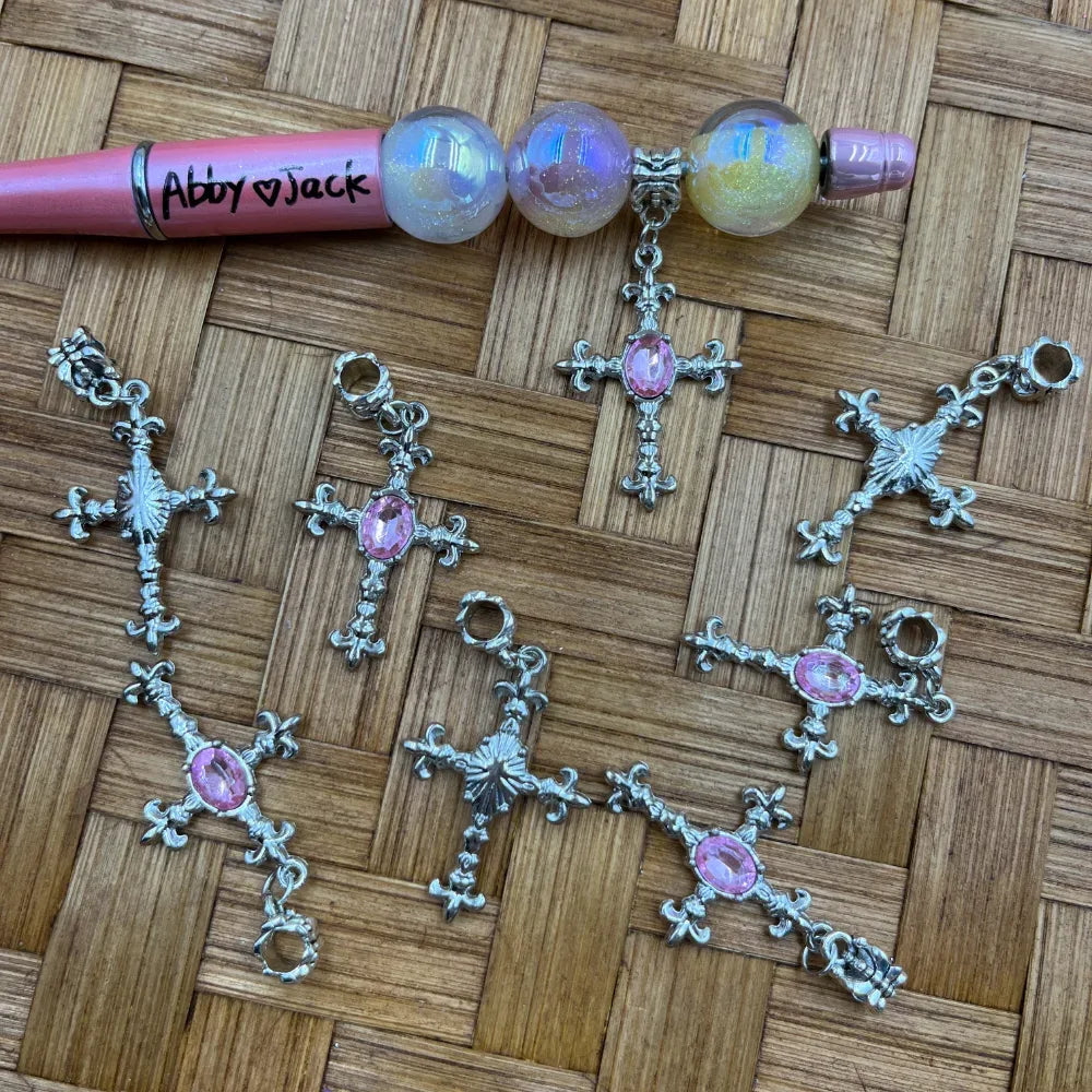 Silver ornate cross charm bail beads with pink oval rhinestone center displayed on wicker surface alongside a pink beadable pen decorated with iridescent glitter globe beads and a cross charm