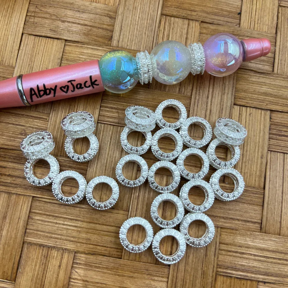 Silver pearl beaded ring metal spacer beads displayed on a woven bamboo surface next to a pink beadable pen decorated with iridescent globe beads and matching spacers, showing the textured circular ring design.