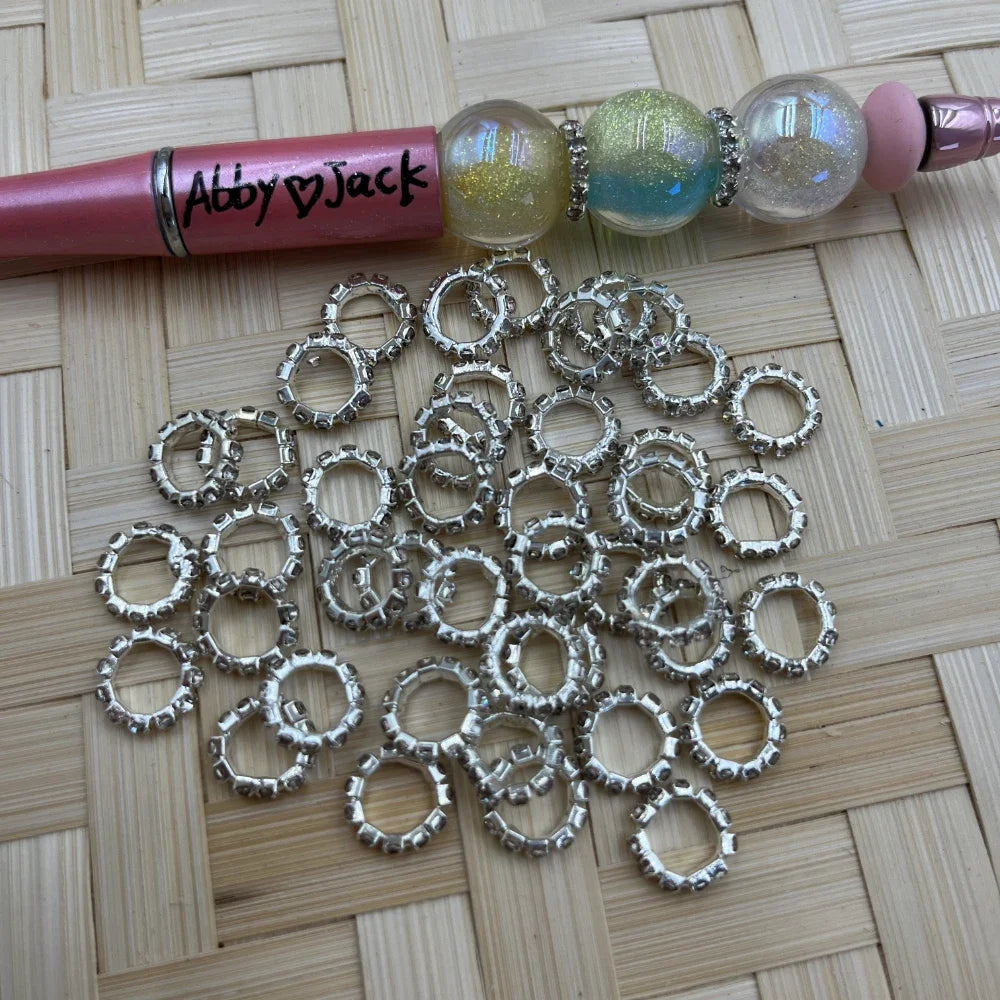 Silver rhinestone ring metal spacer beads scattered on wicker surface next to a pink beadable pen with glitter globe beads separated by matching rhinestone ring spacers.