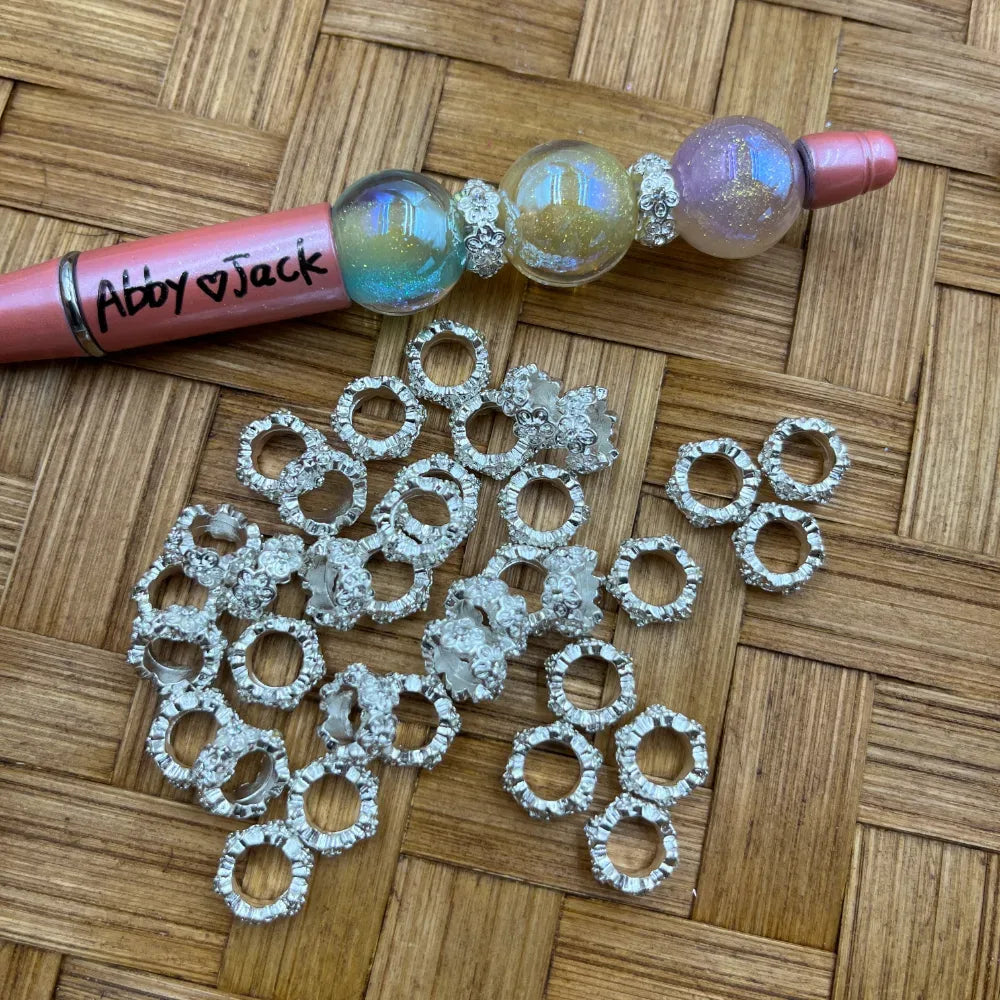 Silver rhinestone ring metal spacer beads with decorative floral edging displayed on wicker surface next to a beadable pen with iridescent galaxy beads