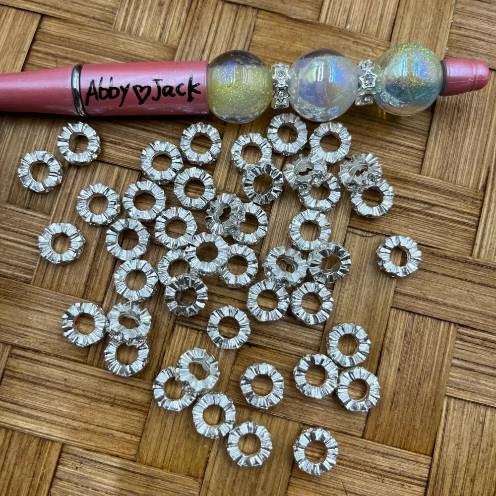 Silver rhinestone rondelle metal spacer beads with serrated crystal-cut edges and open center holes, scattered on wicker surface next to a beadable pen with iridescent globe beads.
