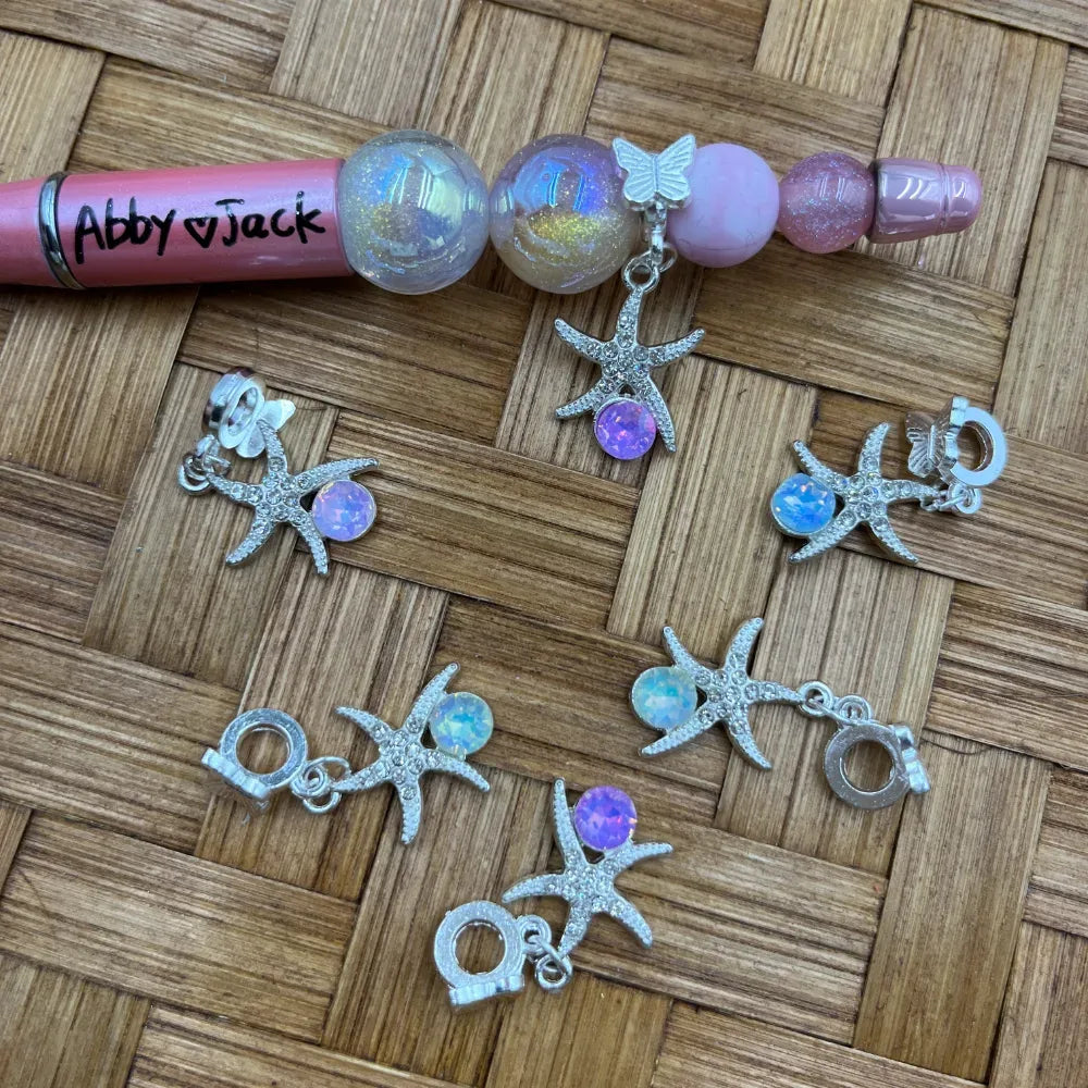 Silver rhinestone starfish charm bail beads with iridescent opal center gemstone, displayed on woven wood surface next to a decorated beadable pen with glitter globe beads labeled Abby and Jack.
