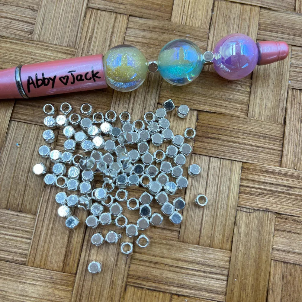 Pile of silver square tube metal spacer beads with wide holes on a wicker surface, shown next to a beadable pen decorated with iridescent globe beads.