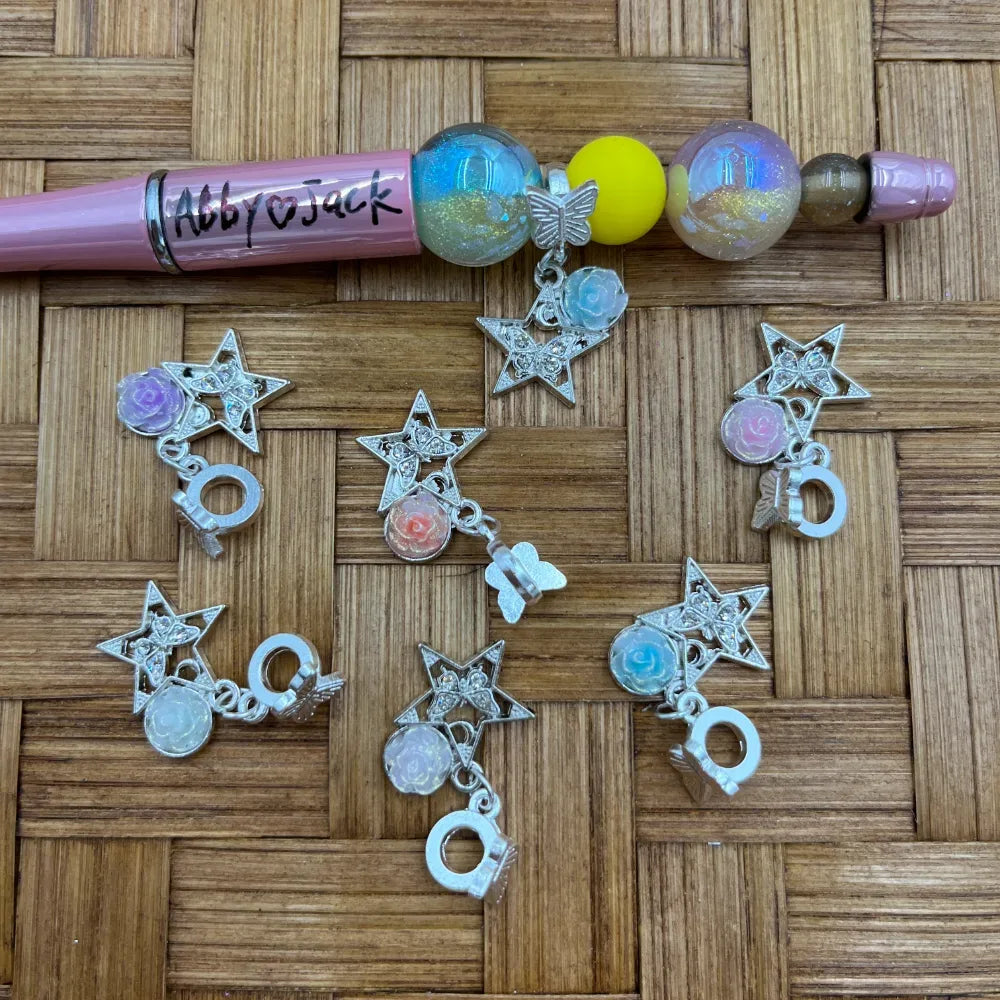 Silver rhinestone star bail beads with pastel rose flower and butterfly charms arranged on wicker surface next to a pink beadable pen labeled Abby and Jack