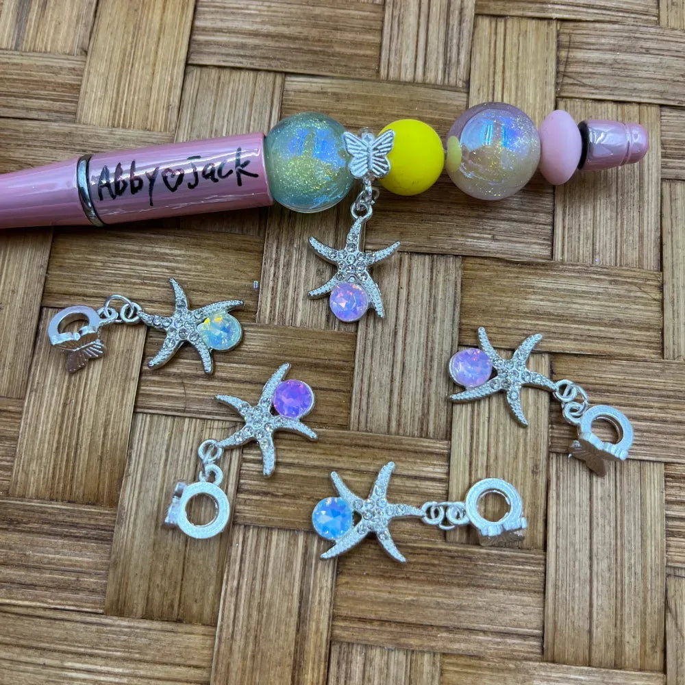 Silver starfish-shaped rhinestone bail beads with iridescent opal gemstone center, shown with a decorated pink beadable pen on a wicker background.