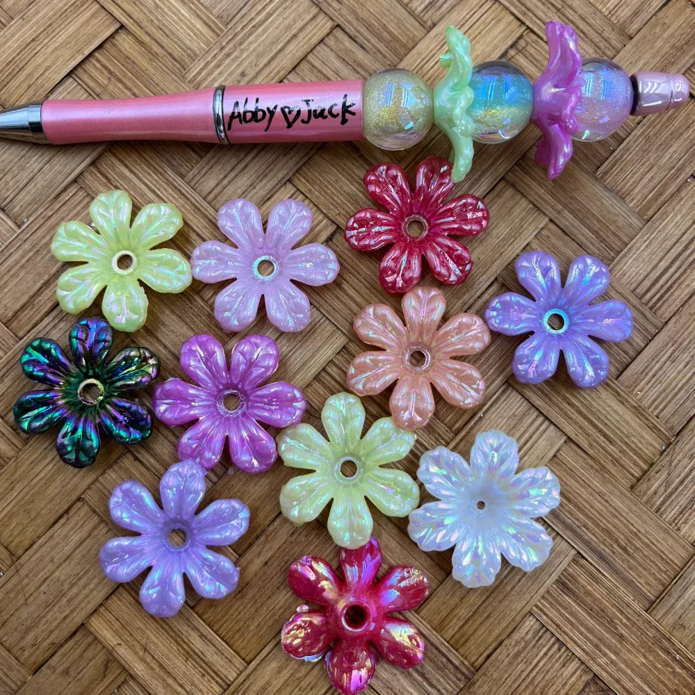 Assorted daisy-shaped AB acrylic spacer beads in pastel and bright iridescent colors displayed beside a pink beadable pen.
