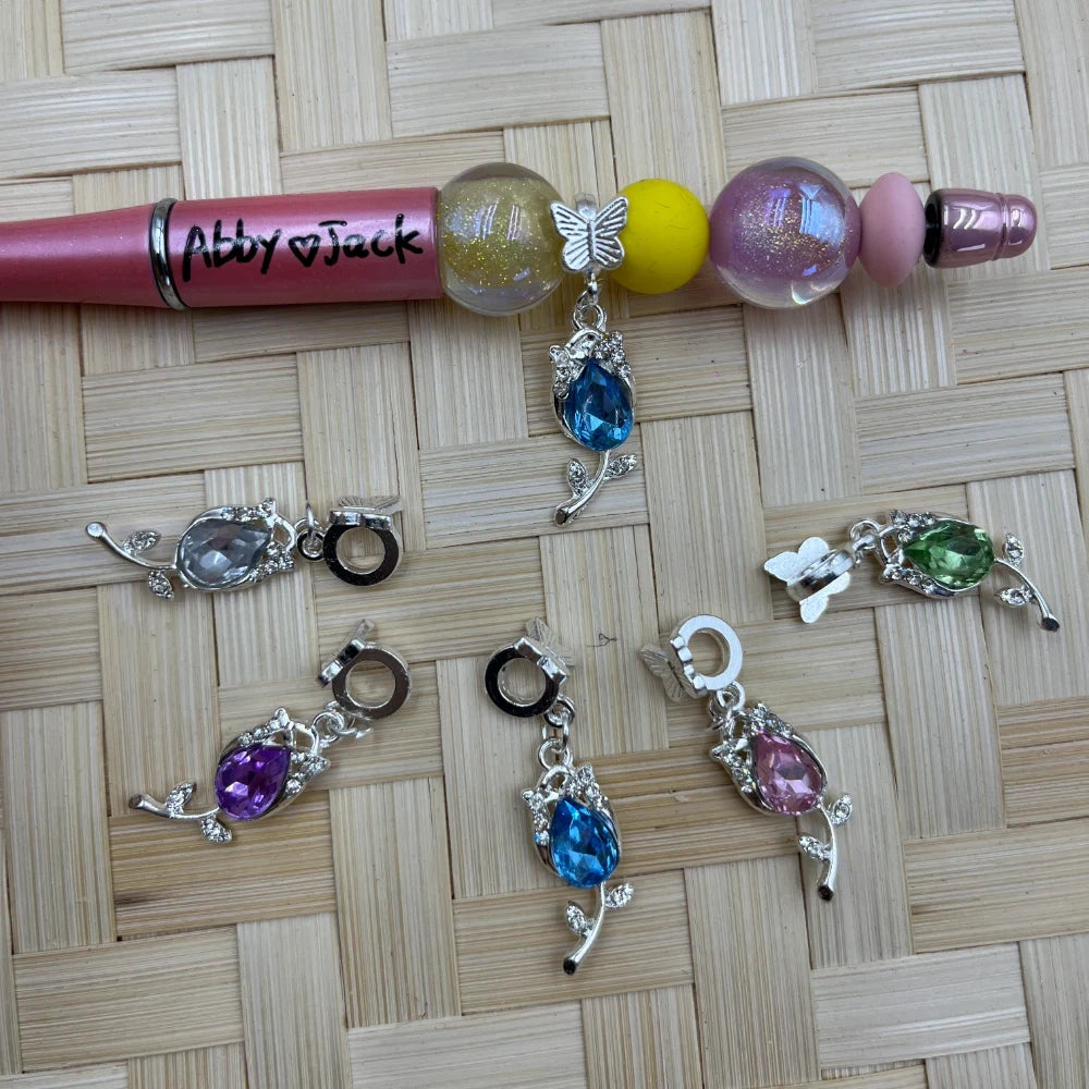 Set of silver-tone floral bail beads with teardrop crystal drops in blue, purple, pink, green, and clear beside a pink beadable pen.