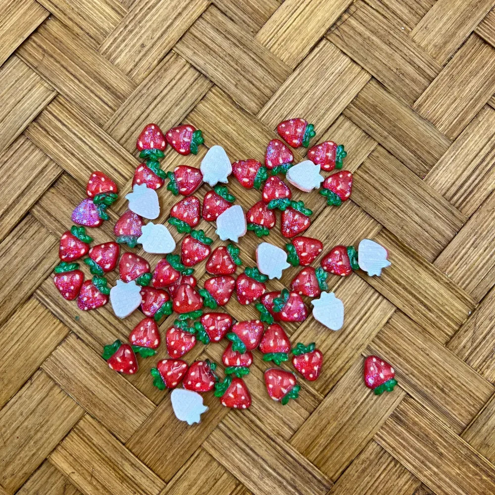 Red strawberry-shaped acrylic beads with white seed dots and green leaf tops scattered on a woven wood surface.