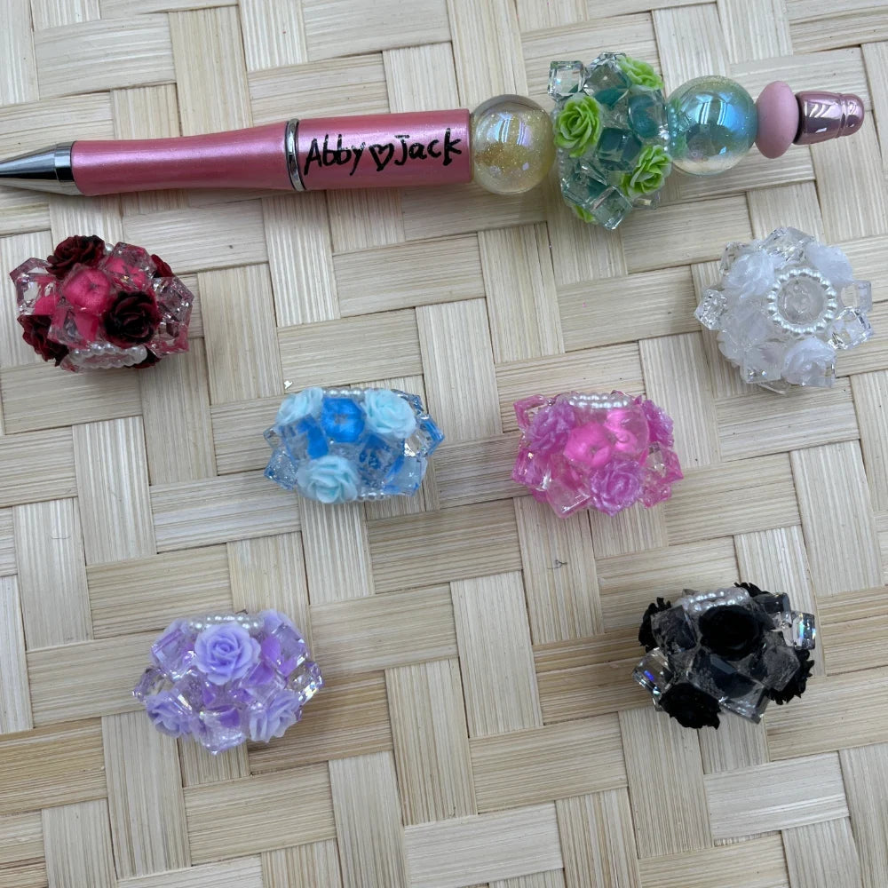 Assorted colorful faceted rose resin fancy beads with clear crystal-look accents displayed beside a pink beadable pen on a woven mat