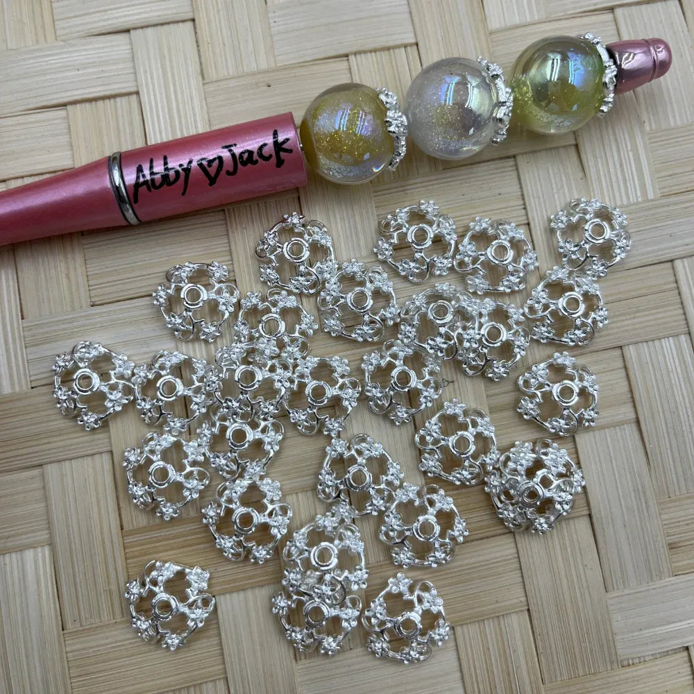Silver floral filigree metal bead caps scattered beside a decorated beadable pen on a woven mat