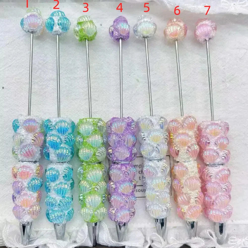 Seven pastel iridescent shell rhinestone pens with shell-shaped bead accents in clear pink, blue, green, purple, white, peach, and pink