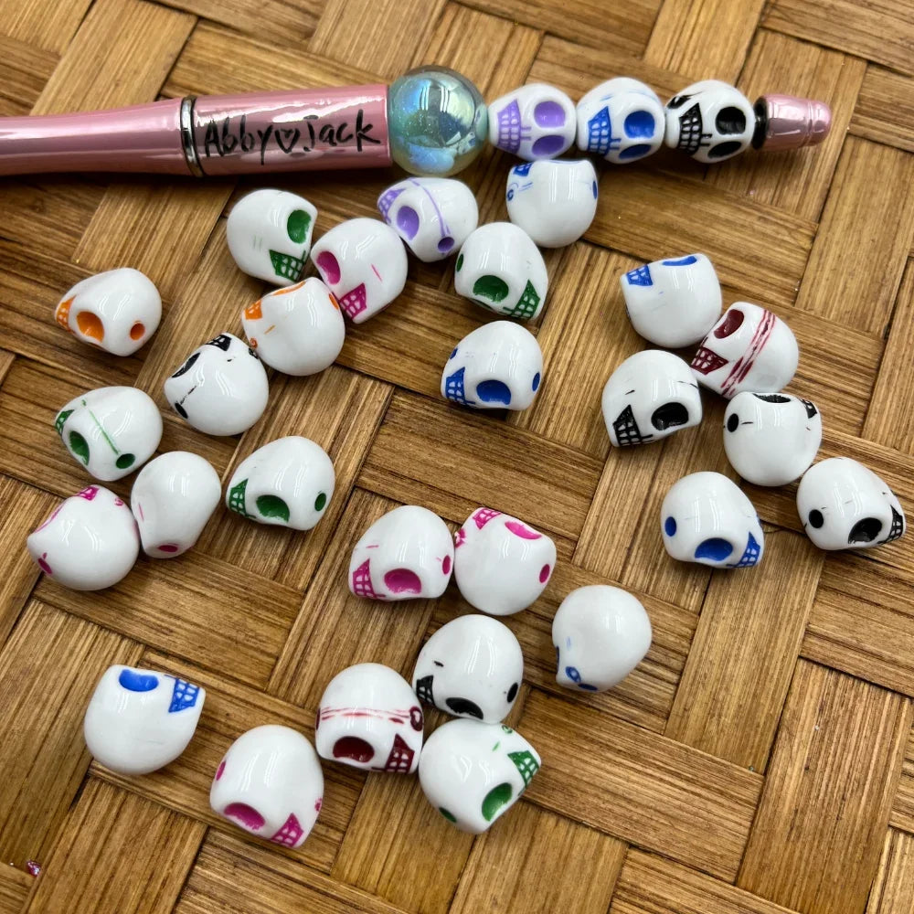 White acrylic skull beads with multicolor painted eye sockets and teeth in orange, green, pink, purple, blue, and black, scattered on a wicker surface next to a pink beadable pen.