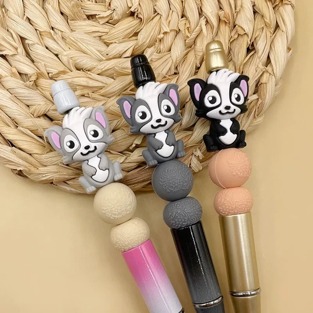Three cute cartoon skunk silicone focal beads in gray, dark gray, and black colorways displayed on beadable pens with decorative silicone beads on a woven mat background.