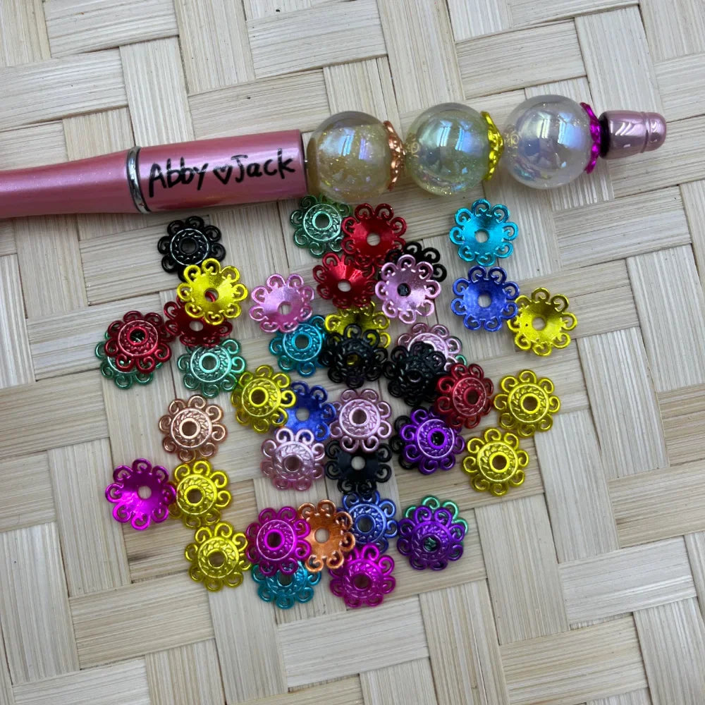 Assorted small multicolor flower alloy bead caps beside a pink beadable pen on a woven background.