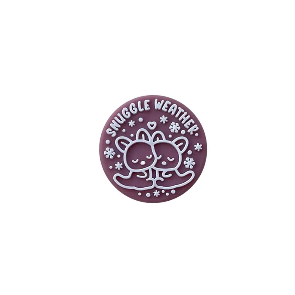 Snuggle Weather Bear silicone focal beads in pink, red, navy blue, dark red, and teal colors displayed on a white ceramic plate and pink surface, featuring a cute cartoon bear hugging design with snowflakes and 'Snuggle Weather' text. - 2