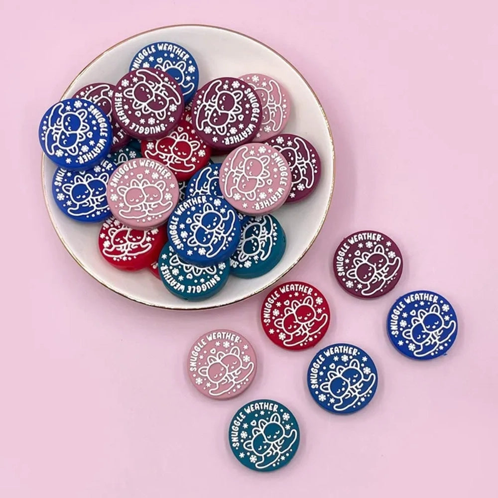 Snuggle Weather Bear silicone focal beads in pink, red, navy blue, dark red, and teal colors displayed on a white ceramic plate and pink surface, featuring a cute cartoon bear hugging design with snowflakes and 'Snuggle Weather' text. - Random Mixed