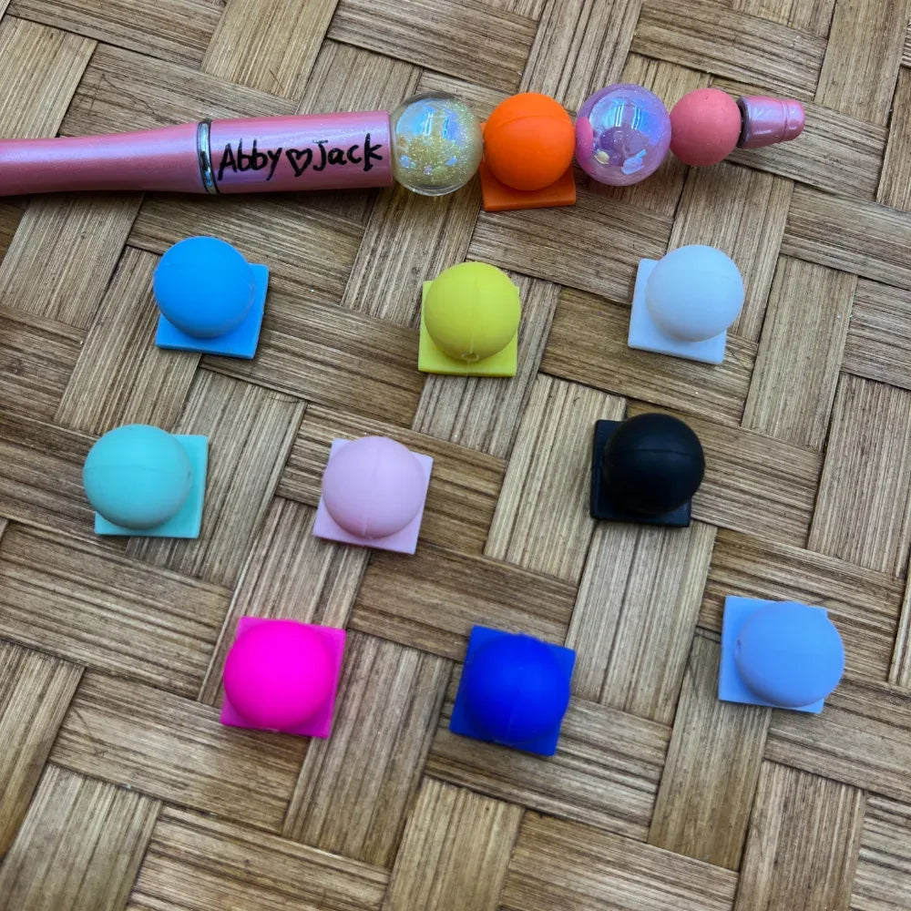 Assorted solid color round silicone beads in blue, teal, yellow, pink, white, black, magenta, cobalt blue, and lavender displayed on a woven surface next to a beadable pen with beads attached, branded Abby and Jack.