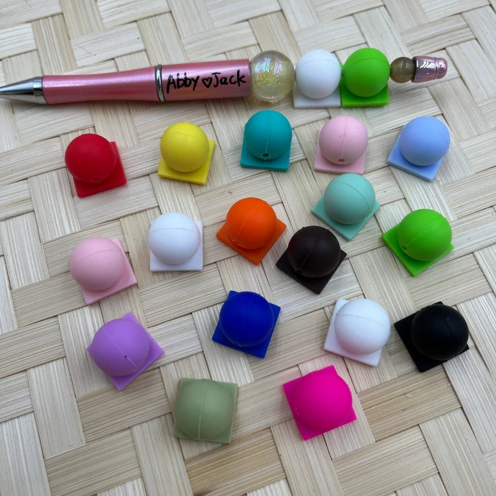 Assorted solid color silicone beads in red, yellow, teal, pink, white, green, orange, brown, blue, purple, black, hot pink, and more, displayed on a wicker surface next to a beadable pen labeled Abby Jack.
