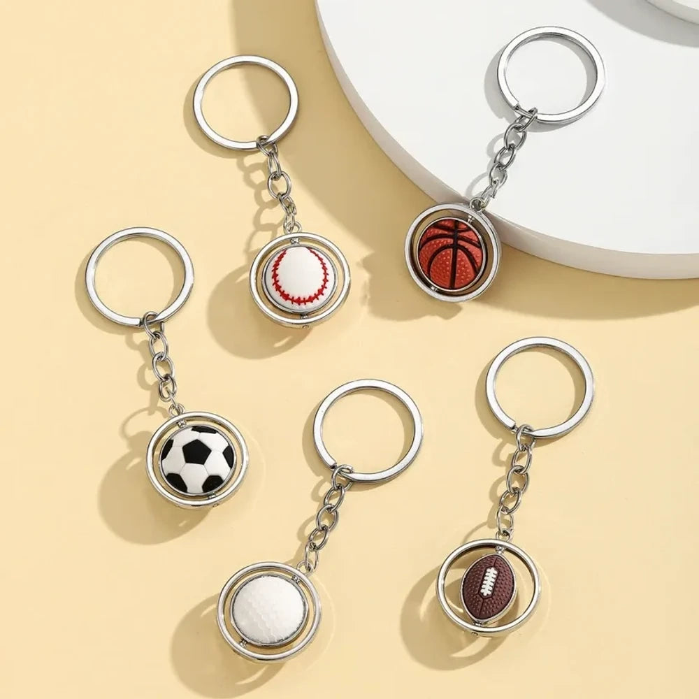 Set of 5 spinning sports ball keychain charms including soccer ball, basketball, football, baseball, and golf ball with silver metal rings on a cream background