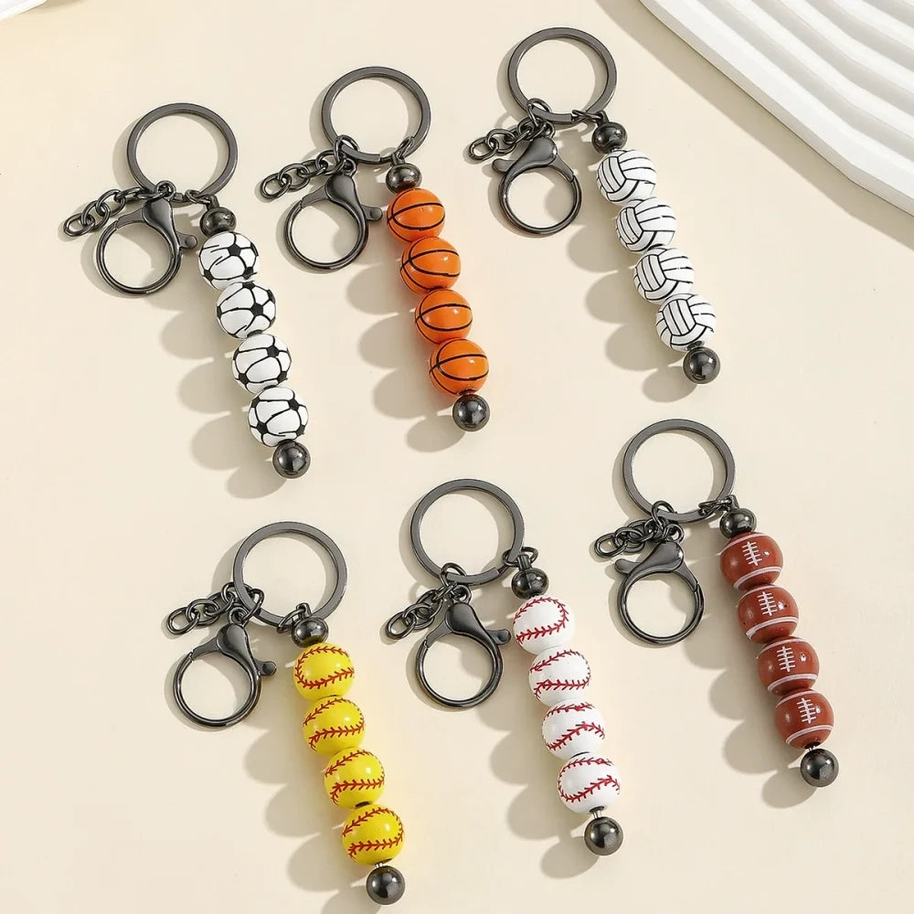 Six sport-themed acrylic bead keychains featuring soccer, basketball, volleyball, softball, baseball, and football ball-shaped beads strung on gunmetal key rings displayed on a beige surface.