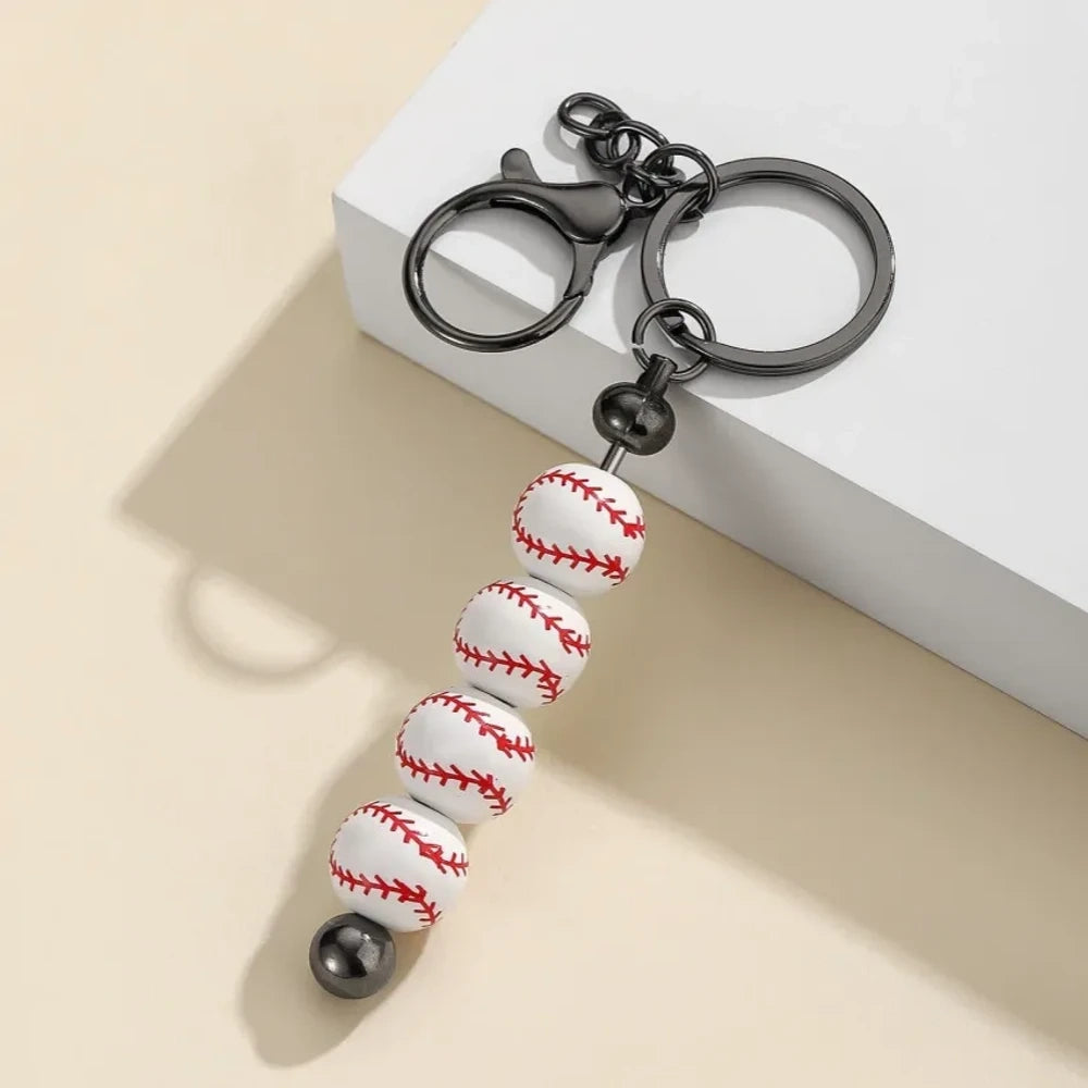 Six sport-themed acrylic bead keychains featuring soccer, basketball, volleyball, softball, baseball, and football ball-shaped beads strung on gunmetal key rings displayed on a beige surface. - Baseball