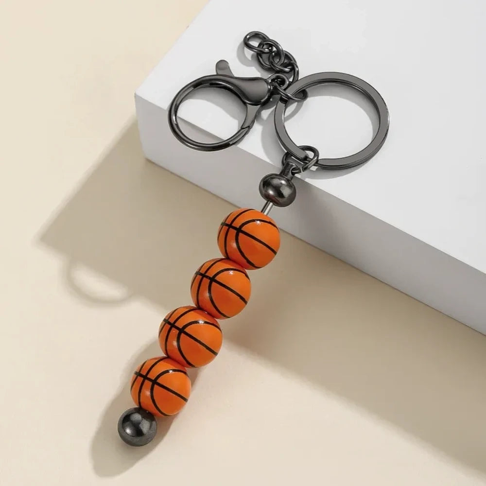 Six sport-themed acrylic bead keychains featuring soccer, basketball, volleyball, softball, baseball, and football ball-shaped beads strung on gunmetal key rings displayed on a beige surface. - Basketball