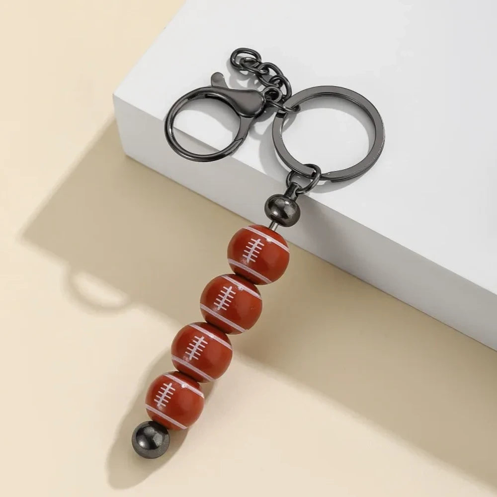 Six sport-themed acrylic bead keychains featuring soccer, basketball, volleyball, softball, baseball, and football ball-shaped beads strung on gunmetal key rings displayed on a beige surface. - Rugby