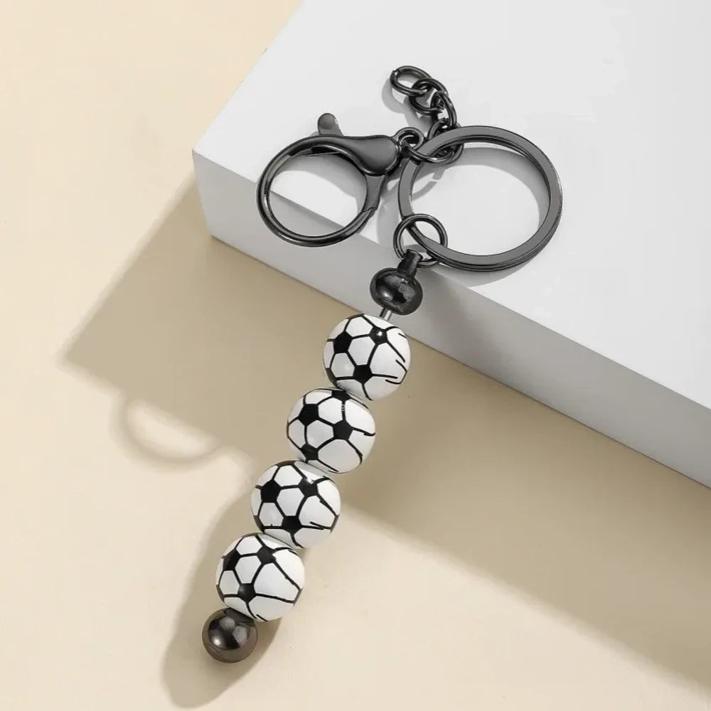 Six sport-themed acrylic bead keychains featuring soccer, basketball, volleyball, softball, baseball, and football ball-shaped beads strung on gunmetal key rings displayed on a beige surface. - Soccer