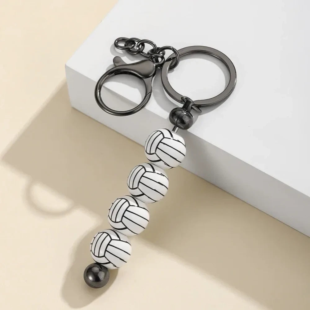 Six sport-themed acrylic bead keychains featuring soccer, basketball, volleyball, softball, baseball, and football ball-shaped beads strung on gunmetal key rings displayed on a beige surface. - Volleyball