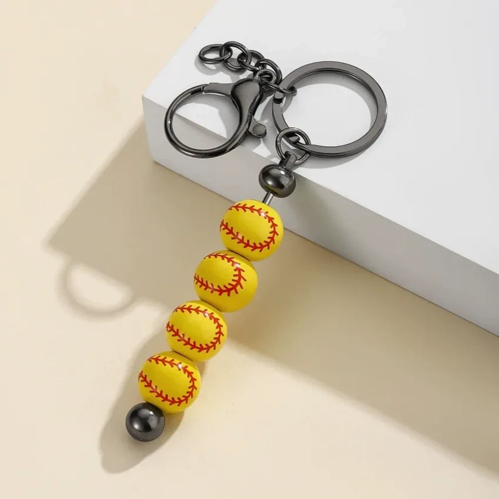 Six sport-themed acrylic bead keychains featuring soccer, basketball, volleyball, softball, baseball, and football ball-shaped beads strung on gunmetal key rings displayed on a beige surface. - Yellow Baseball