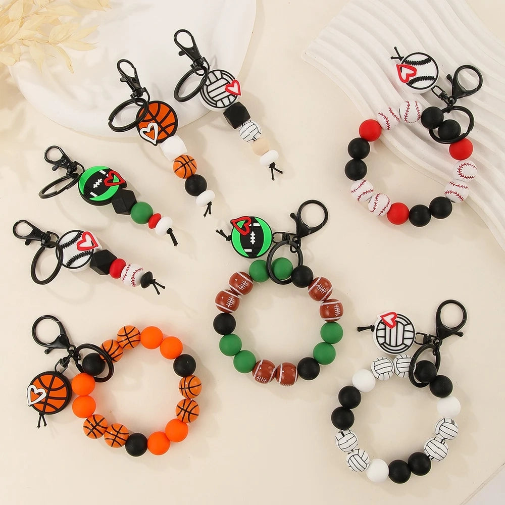 Assorted sports ball printed silicone beads and bracelet keychains featuring basketball, football, baseball, and volleyball designs with black hardware clasps on a white surface.
