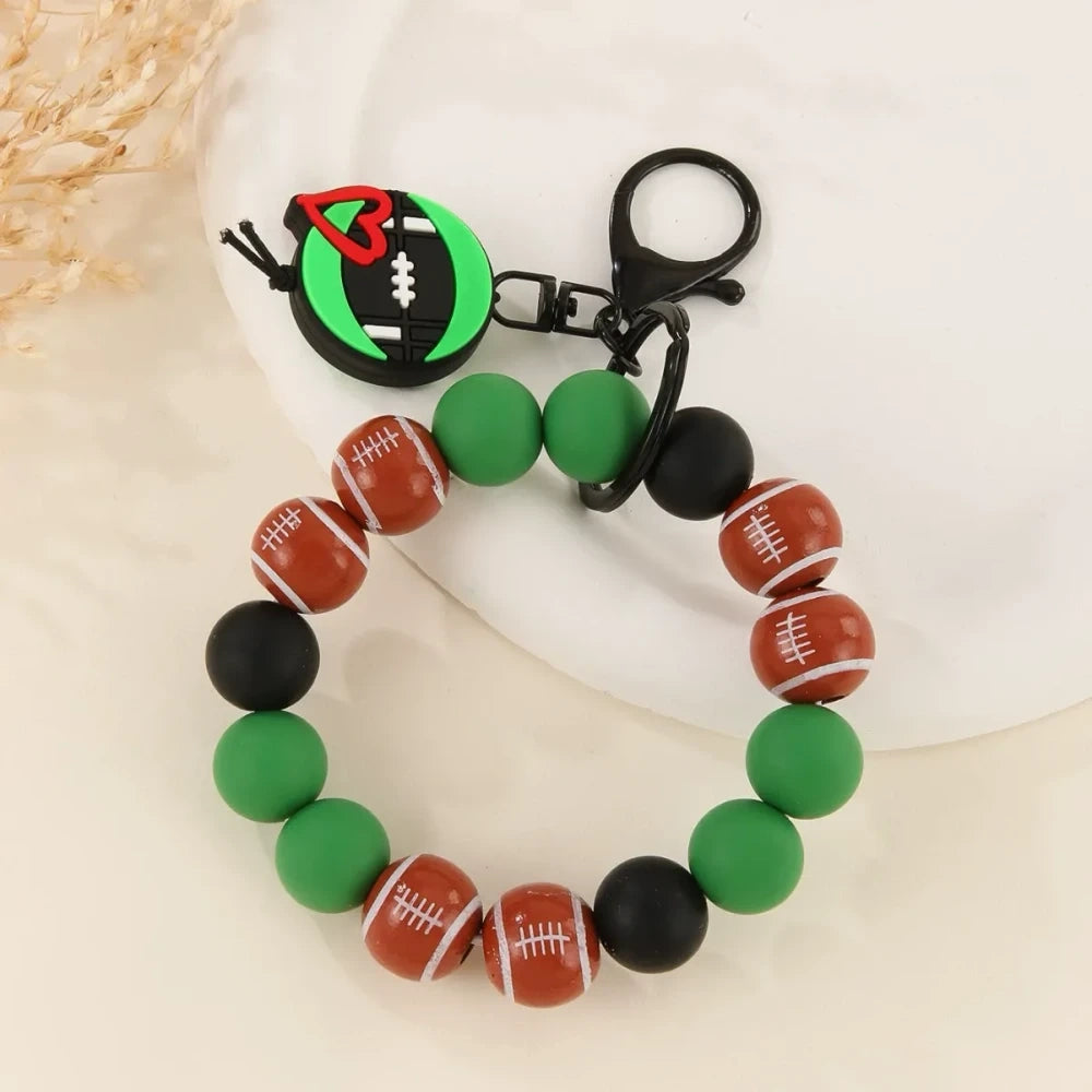 Assorted sports ball printed silicone beads and bracelet keychains featuring basketball, football, baseball, and volleyball designs with black hardware clasps on a white surface. - A