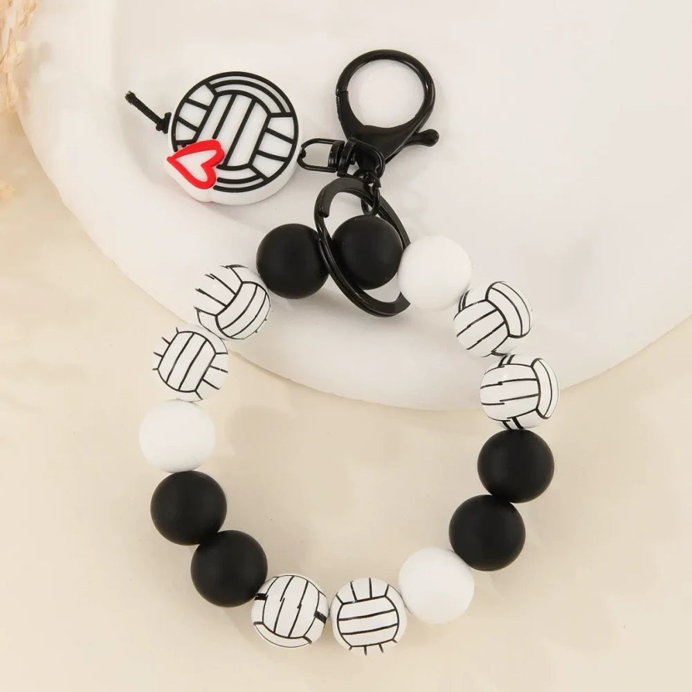 Assorted sports ball printed silicone beads and bracelet keychains featuring basketball, football, baseball, and volleyball designs with black hardware clasps on a white surface. - B