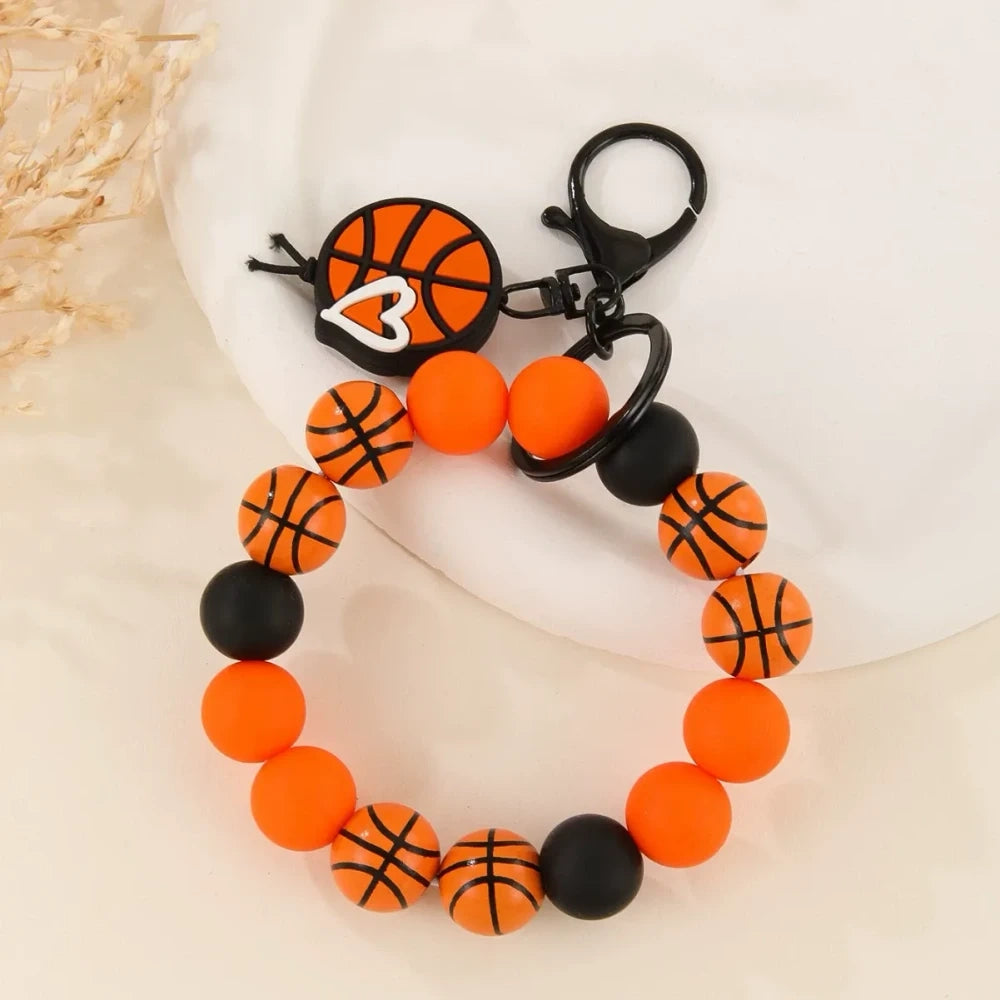 Assorted sports ball printed silicone beads and bracelet keychains featuring basketball, football, baseball, and volleyball designs with black hardware clasps on a white surface. - C