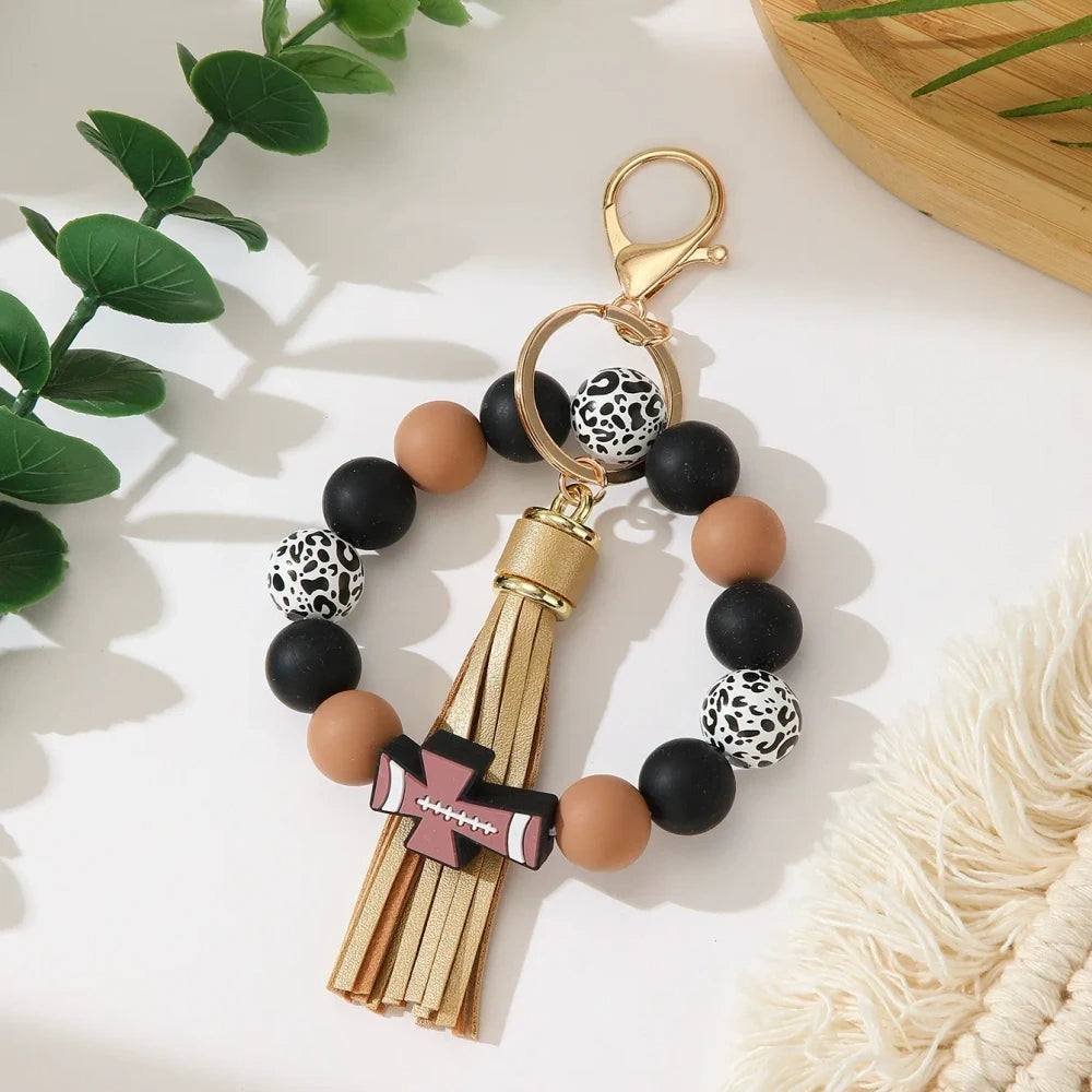 Set of 6 silicone bead stretch bracelet keychains featuring sports cross charms and leather tassels in assorted colors including red, blue, green, orange, black and white with leopard print beads and gold lobster clasp hardware. - Brown