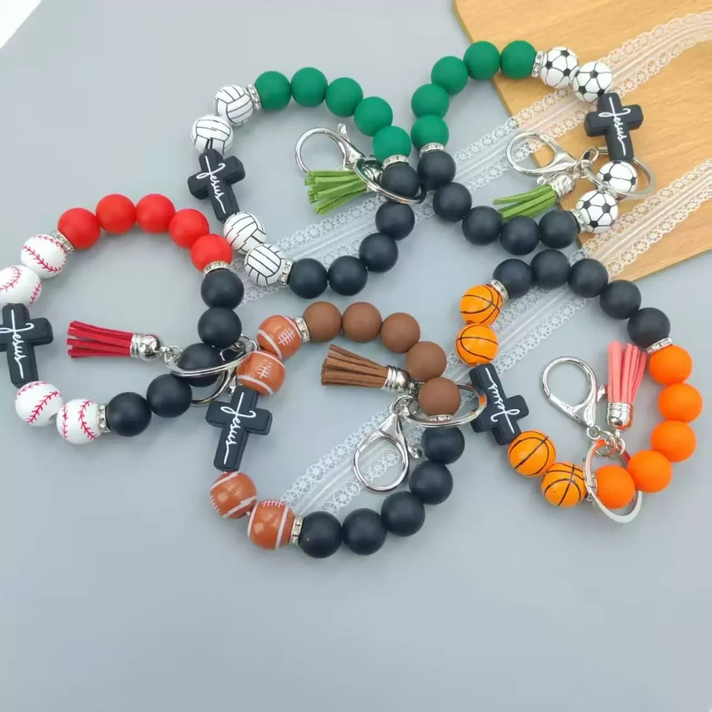 Assorted sports print silicone beads in basketball, football, baseball, soccer, and volleyball designs with matching solid color beads, shown on completed beaded bracelet keychains with cross charms and tassels.