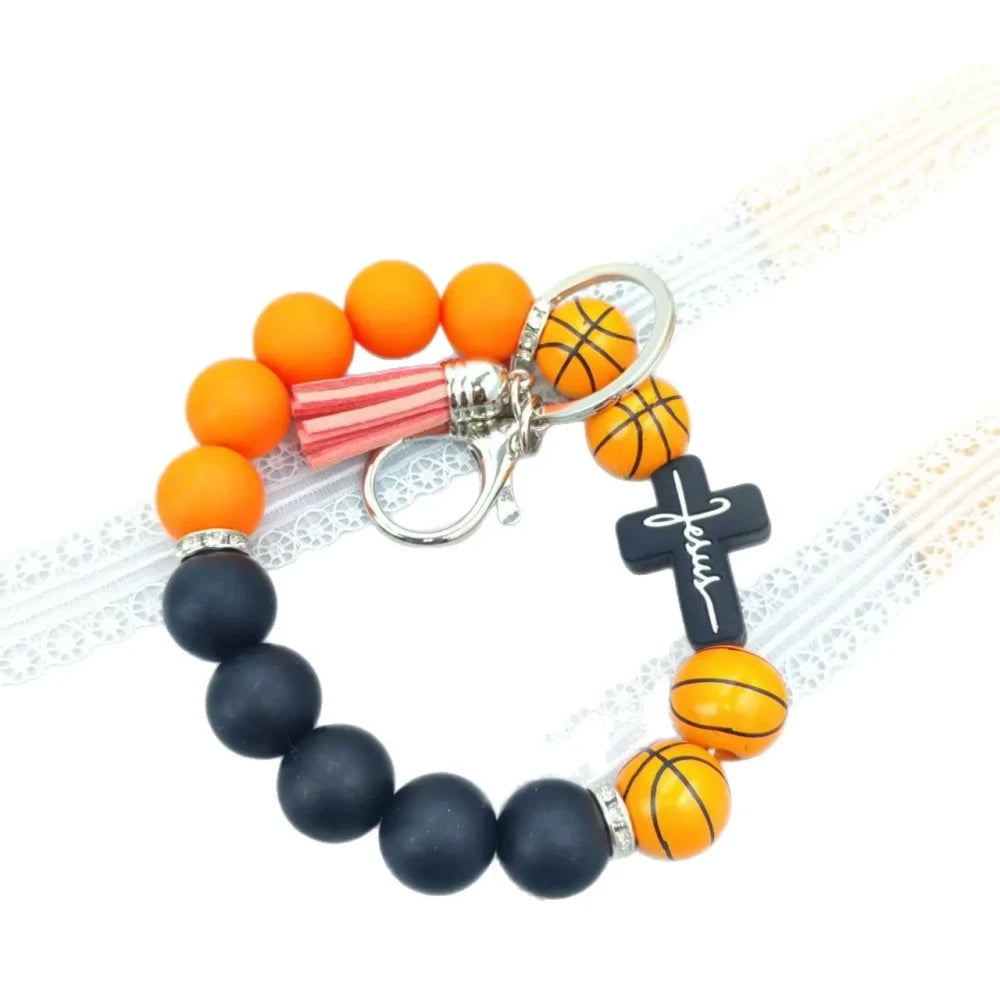 Assorted sports print silicone beads in basketball, football, baseball, soccer, and volleyball designs with matching solid color beads, shown on completed beaded bracelet keychains with cross charms and tassels. - Basketball