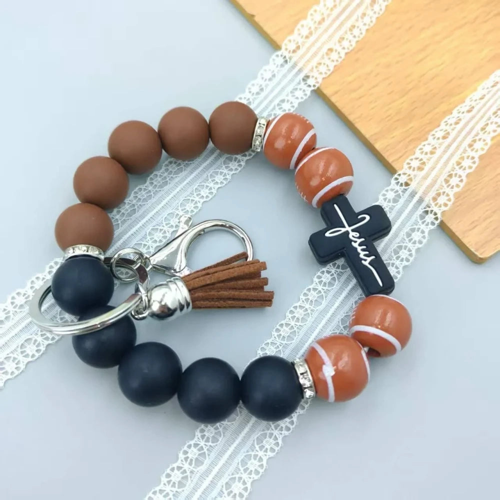Assorted sports print silicone beads in basketball, football, baseball, soccer, and volleyball designs with matching solid color beads, shown on completed beaded bracelet keychains with cross charms and tassels. - Rugby