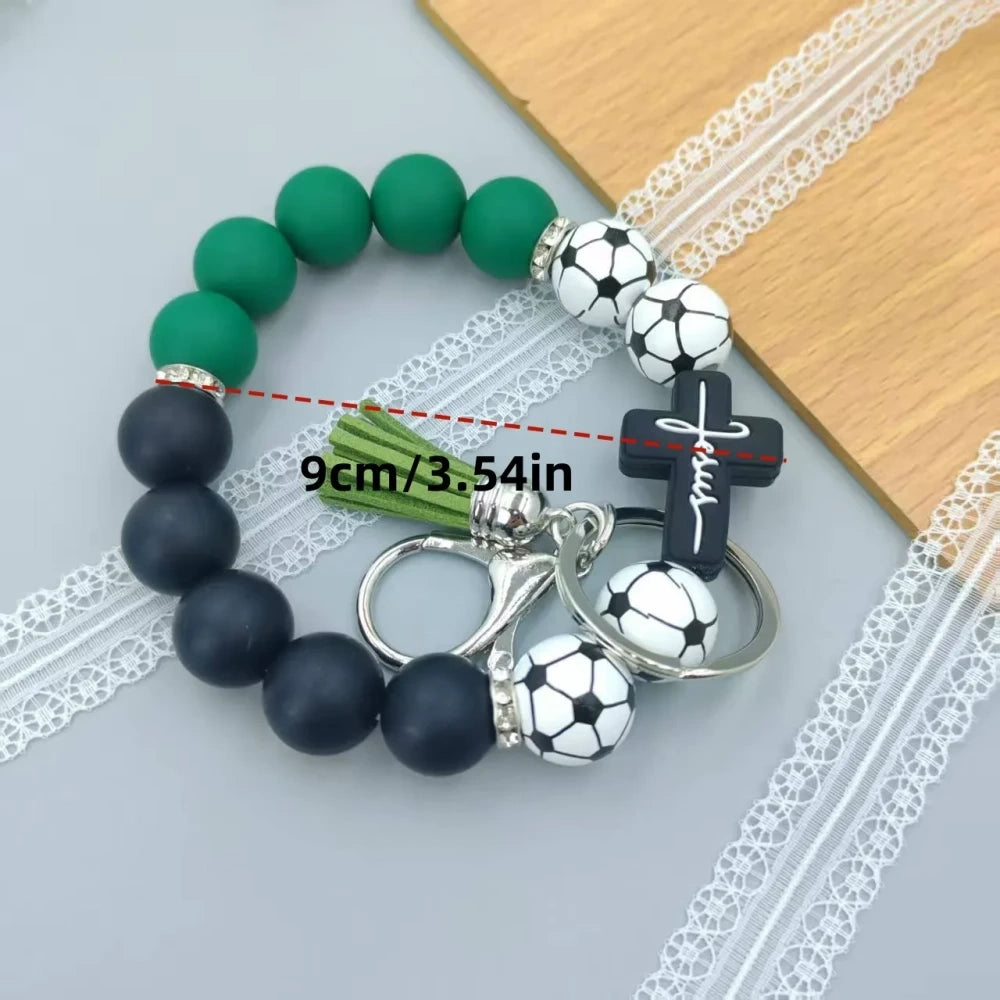 Assorted sports print silicone beads in basketball, football, baseball, soccer, and volleyball designs with matching solid color beads, shown on completed beaded bracelet keychains with cross charms and tassels. - Soccer